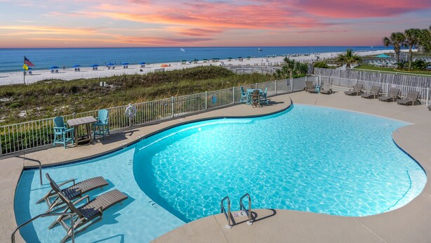 A swimming pool with lounge chairs and a view of the beach at sunset.