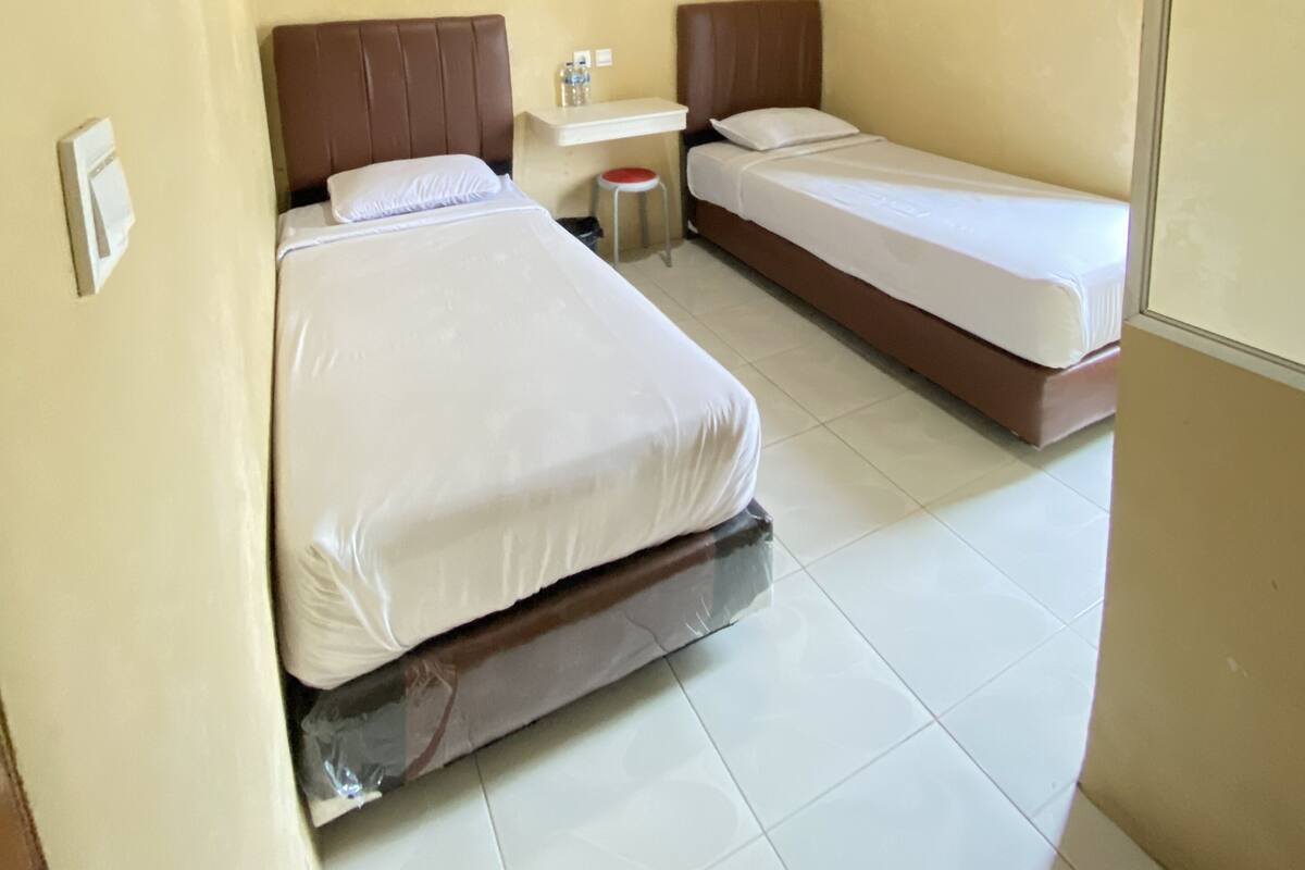 Une chambre d’hôtel avec deux lits simples, une petite table, une chaise et un luminaire fixé au mur.