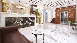 A hotel reception area with a marble floor, a reception desk with a sign reading 'The Dwelling,' a clock, and a staircase leading to an elevator. - Dwelling residency (Greater Noida)