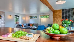 Una sala de estar moderna con una mesa de comedor de madera dispuesta con frutas y quesos, un sofá y vistas a zonas verdes a través de amplios ventanales.