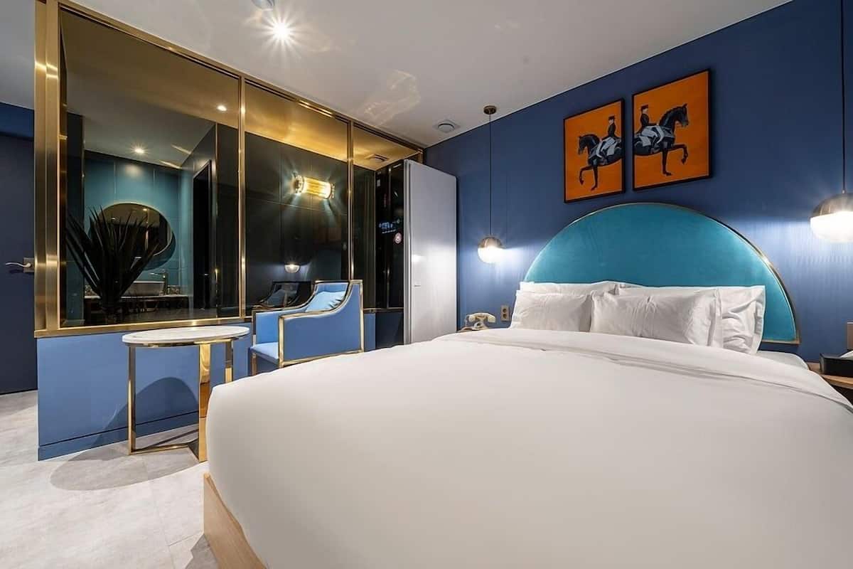 Una habitación de hotel moderna con una cama grande, un área de estar con una mesa y sillas, un espejo amplio y dos obras de arte en la pared.