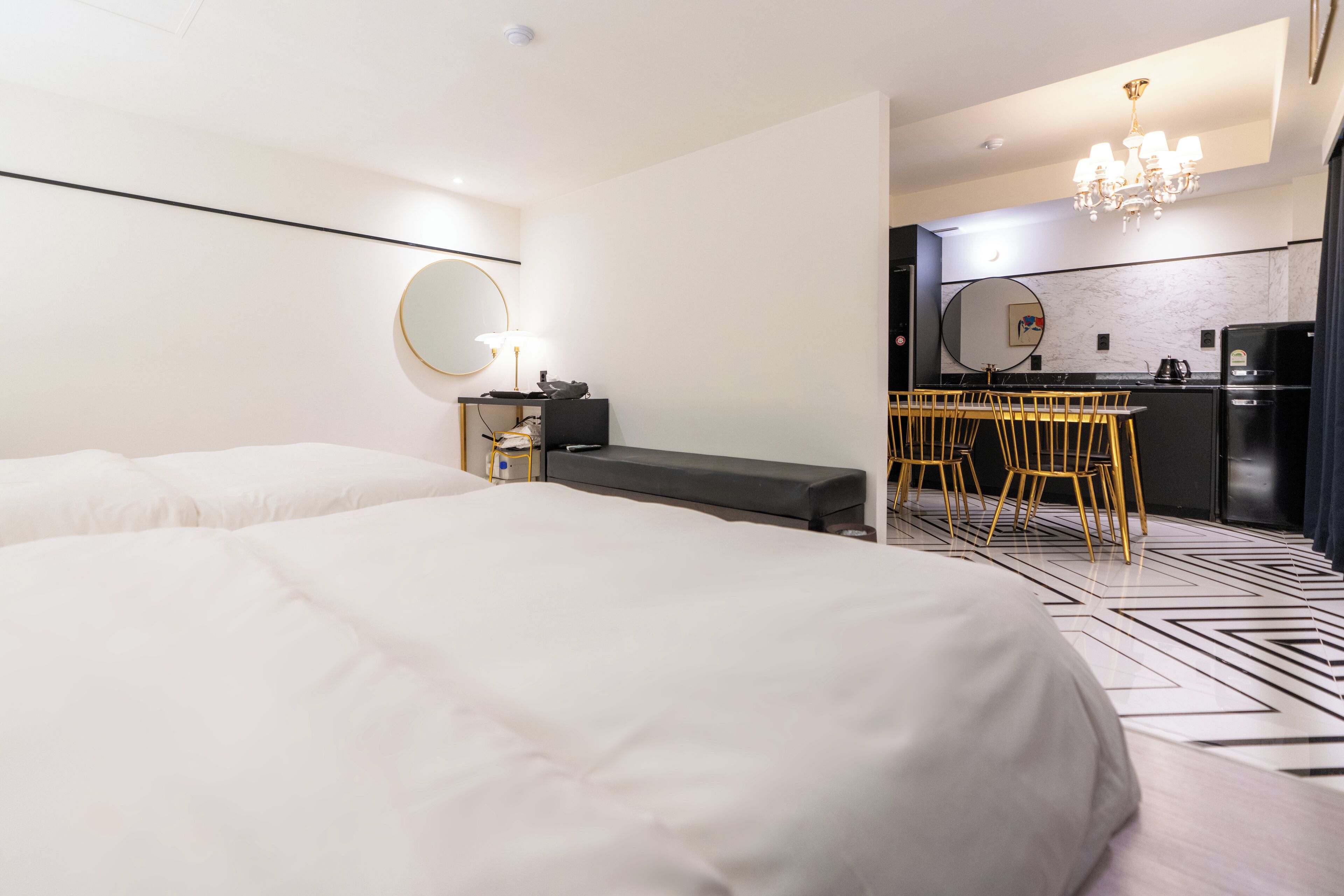 Quarto de hotel moderno com uma cama grande, uma escrivaninha com cadeira, um espelho redondo e uma cozinha compacta.