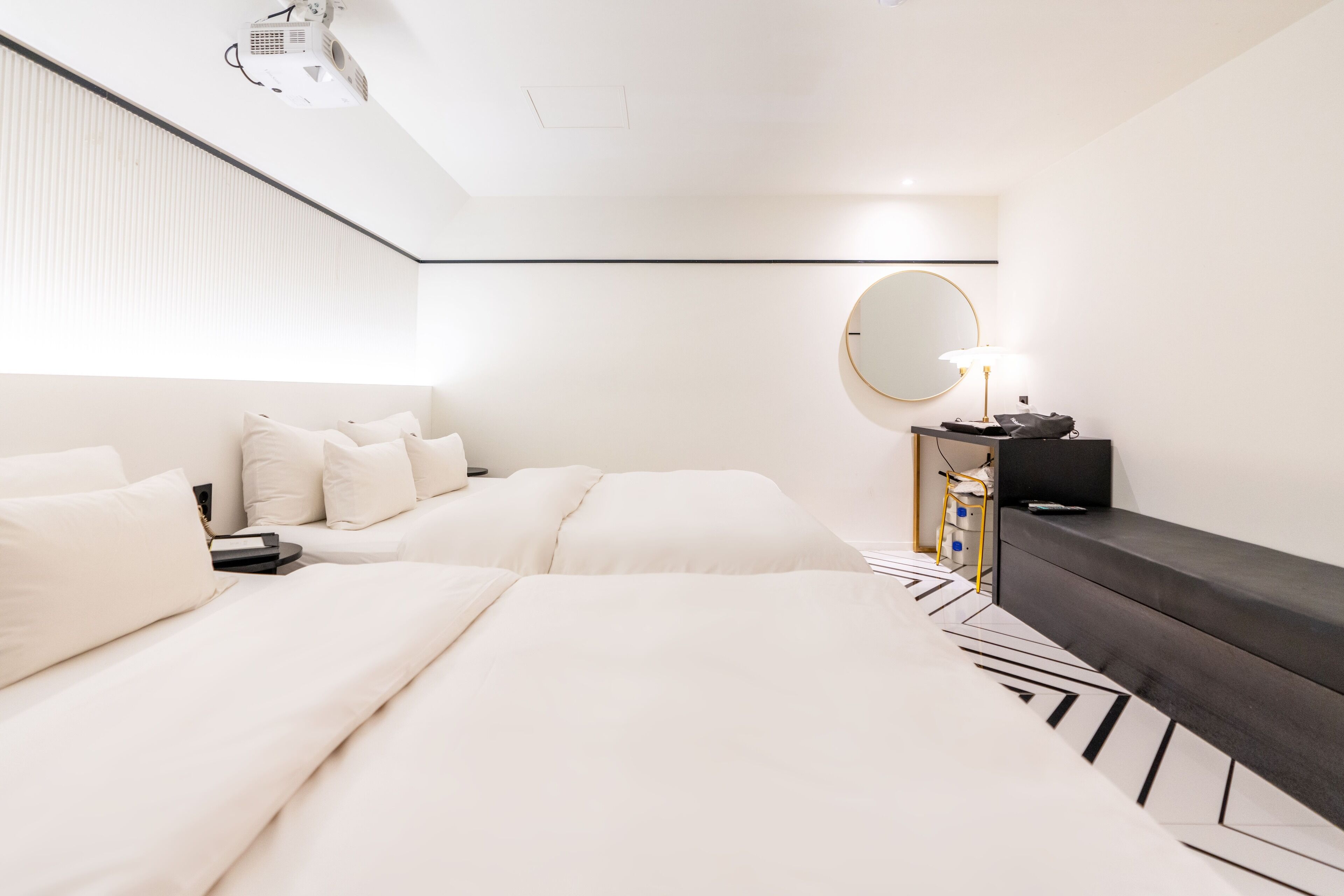 Een moderne hotelkamer met twee bedden, een grote spiegel, een bureau met lamp en een projector aan het plafond.
