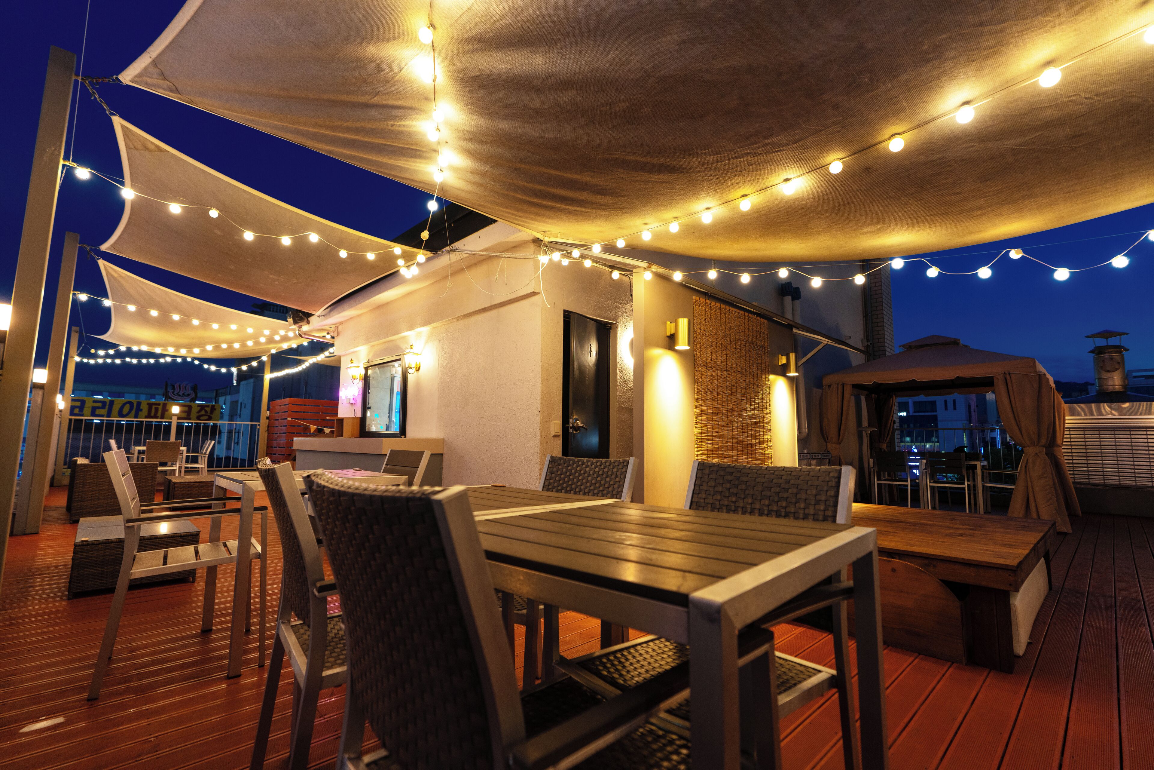 An outdoor dining area with wooden tables and chairs, string lights, and a canopy providing shade.