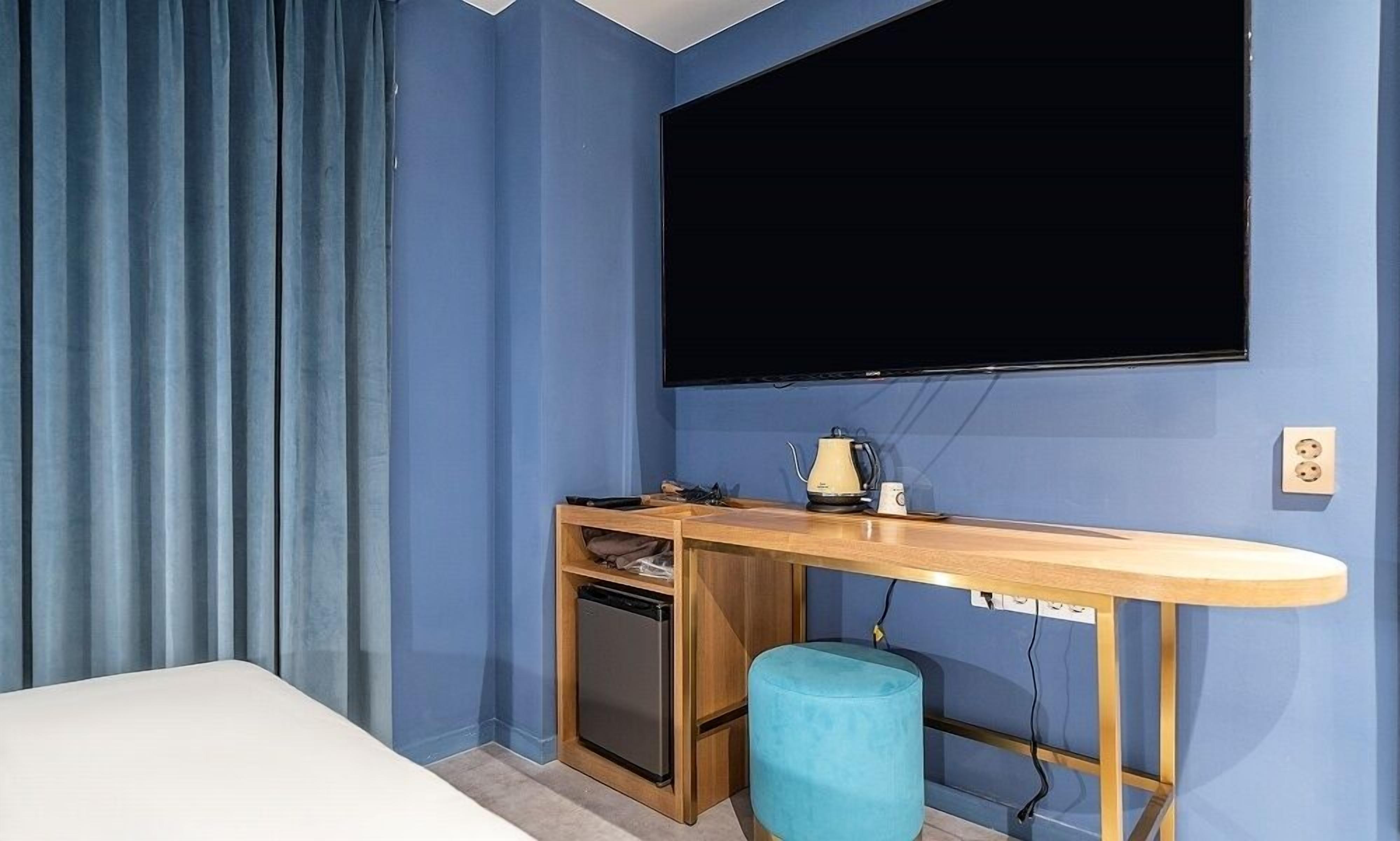 Habitación compacta con cama, escritorio con computadora, refrigerador pequeño, reposapiés azul y televisor montado.