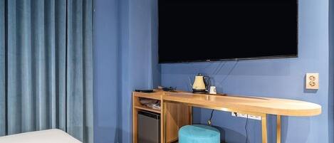 Habitación compacta con cama, escritorio con computadora, refrigerador pequeño, reposapiés azul y televisor montado.
