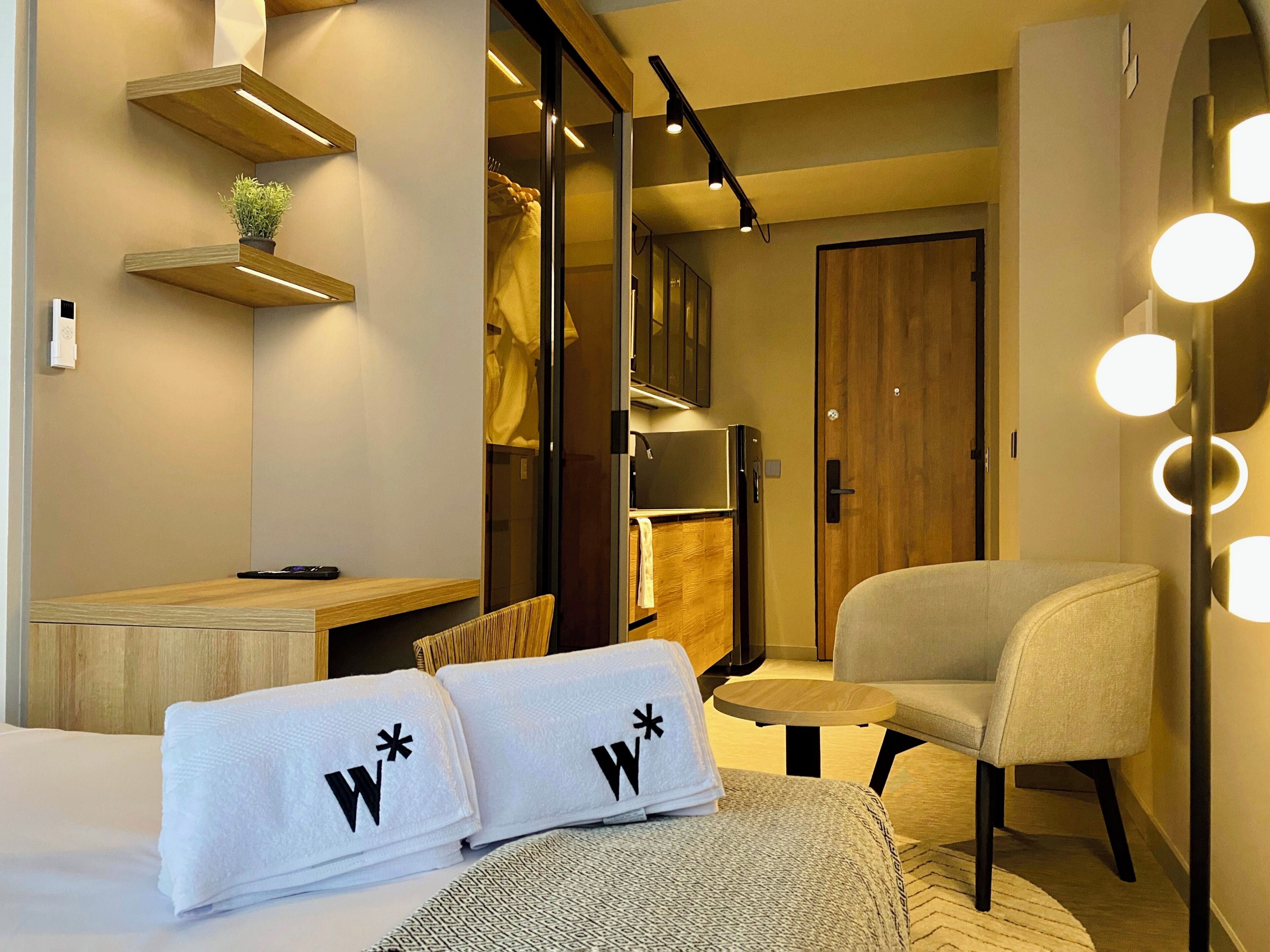A hotel room with a bed, a chair, a desk, a wardrobe, and a mirror with lights.