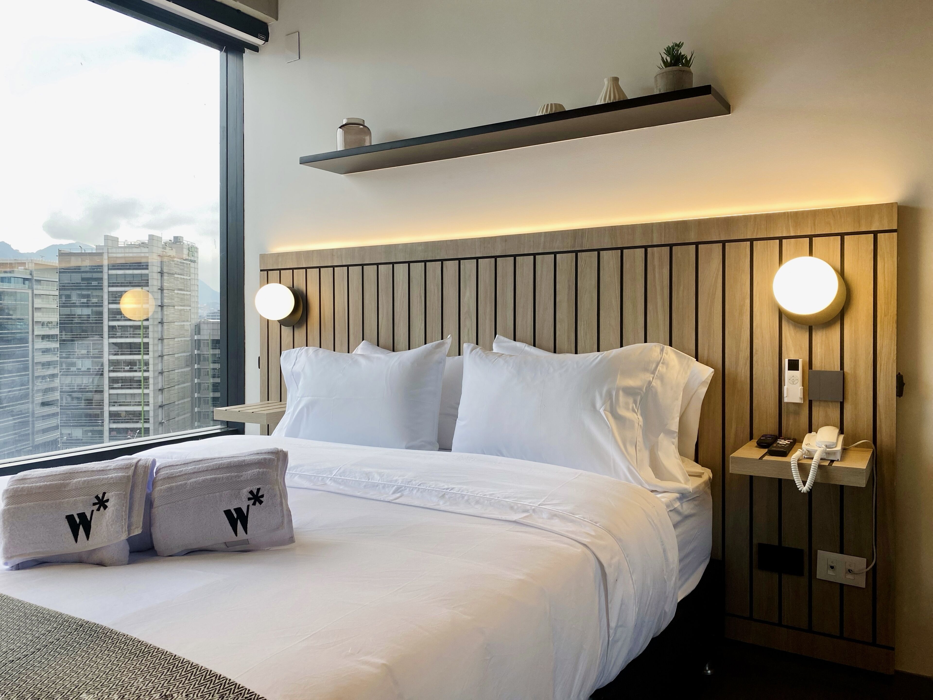 Quarto de hotel com uma cama grande, duas mesas de cabeceira, um telefone fixado na parede e vista para prédios da cidade.