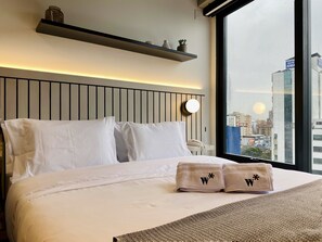 A hotel room with a bed, white bedding, a bedside table with a lamp, a window with a city view, and a shelf with decorative items. - Wynwood House 100 (Bogotá)