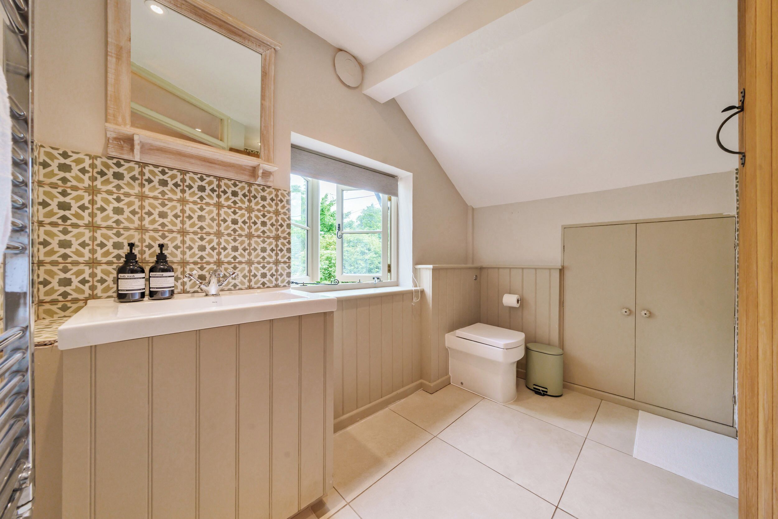 A bathroom with a window, a toilet, a sink with bottles, and a cabinet.