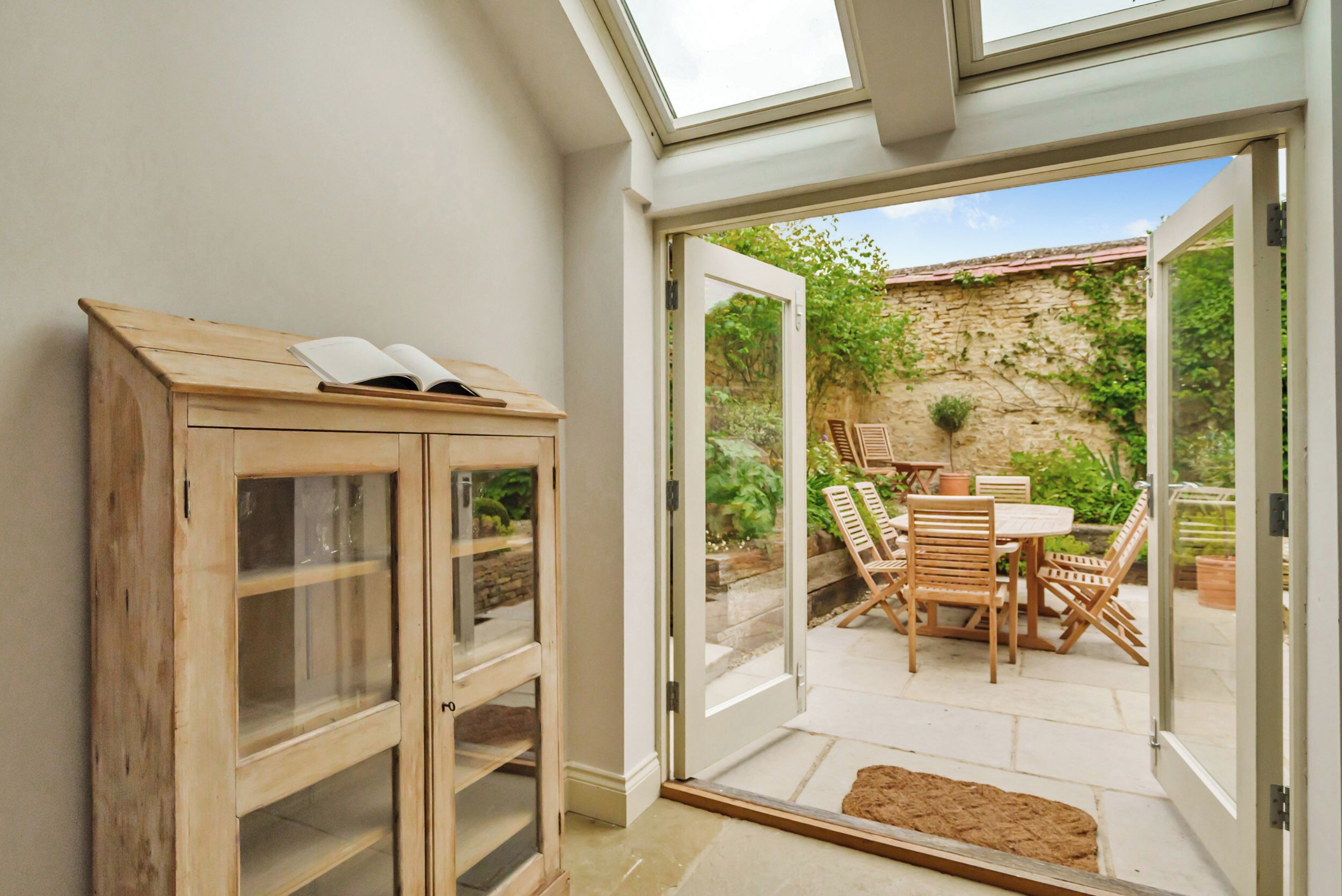A wooden cabinet with glass doors and a book on top, a sliding glass door leading to an outdoor patio with a stone wall, a table, and chairs.
