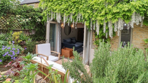 A cozy outdoor seating area with a table and chairs, surrounded by lush greenery and flowering plants. - Lynton Cottage (Chipping Norton)