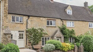 A stone house with a traditional design, featuring a stone facade, multiple windows, and a well-maintained garden with various shrubs and a bench. - Lynton Cottage (Chipping Norton)