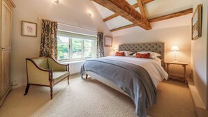 A bedroom with a large bed, a chair, a window with curtains, and wooden beams on the ceiling. - Lynton Cottage (Chipping Norton)