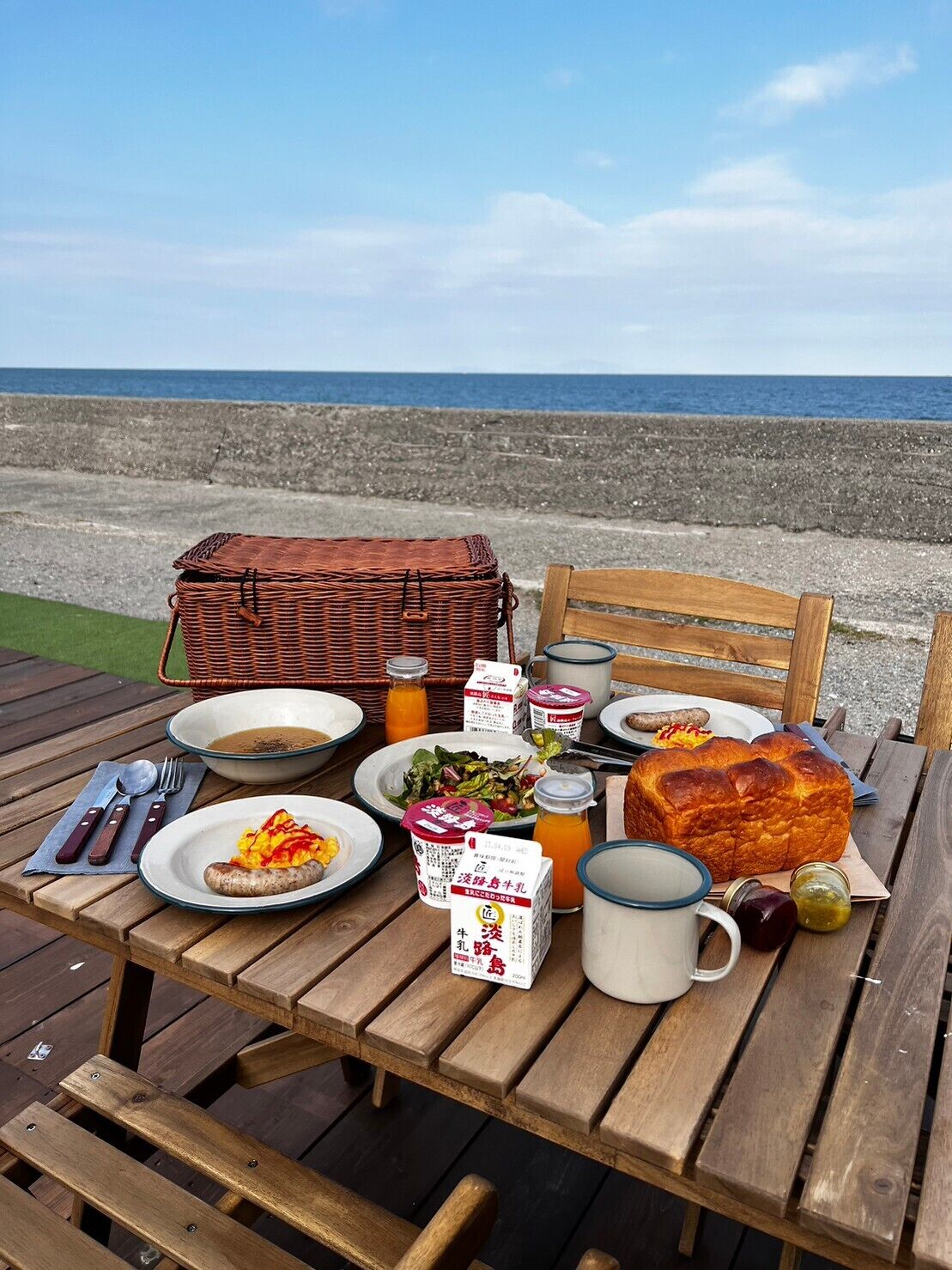 Una mesa de madera dispuesta con comida, incluyendo pan, ensalada y una salchicha, con una cesta de mimbre y una silla de madera al fondo.