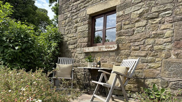 A stone building with a window and two wooden chairs with cushions. - Old Post Office Barn (Leek)