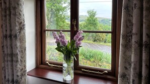 A window with a floral curtain, a vase with flowers, and a view of greenery and a road. - Old Post Office Barn (Leek)
