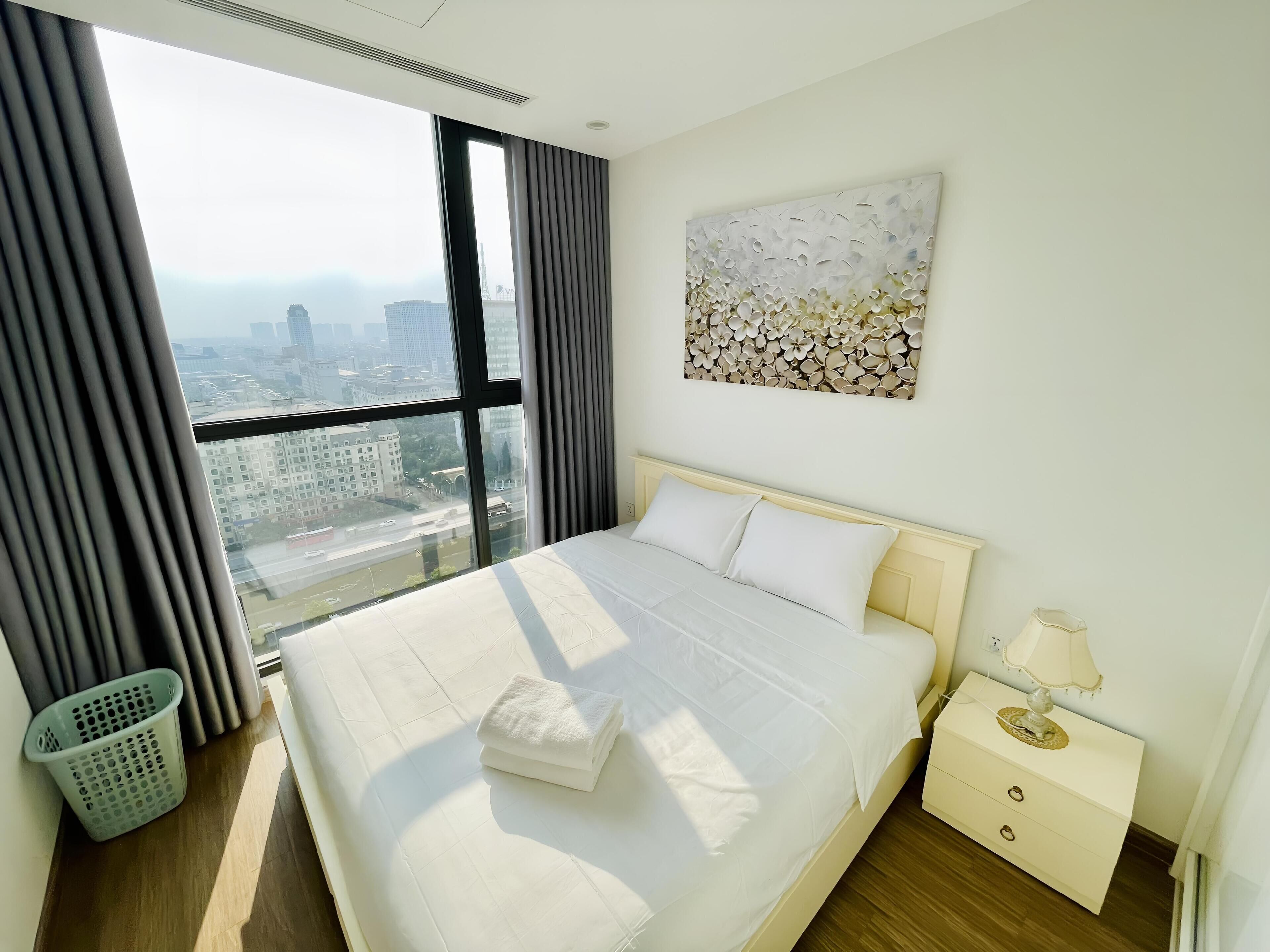 A hotel room with a large bed, a nightstand, a lamp, a wall art piece, and a view of the city through large windows.