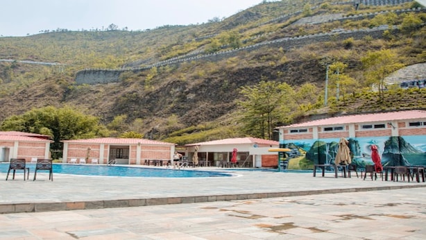 A swimming pool area with a view of a hilly landscape, a mural, and a building with a red roof. - MeroStay 259 KB Resort (Purano Jhangajholi)