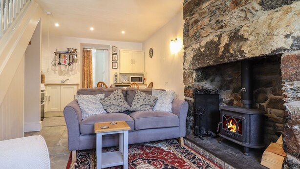 Un salon chaleureux avec un canapé gris, des coussins à motifs, une petite table d’appoint en bois, un tapis rouge et beige à motifs, une cheminée en pierre et, au fond, une cuisine.