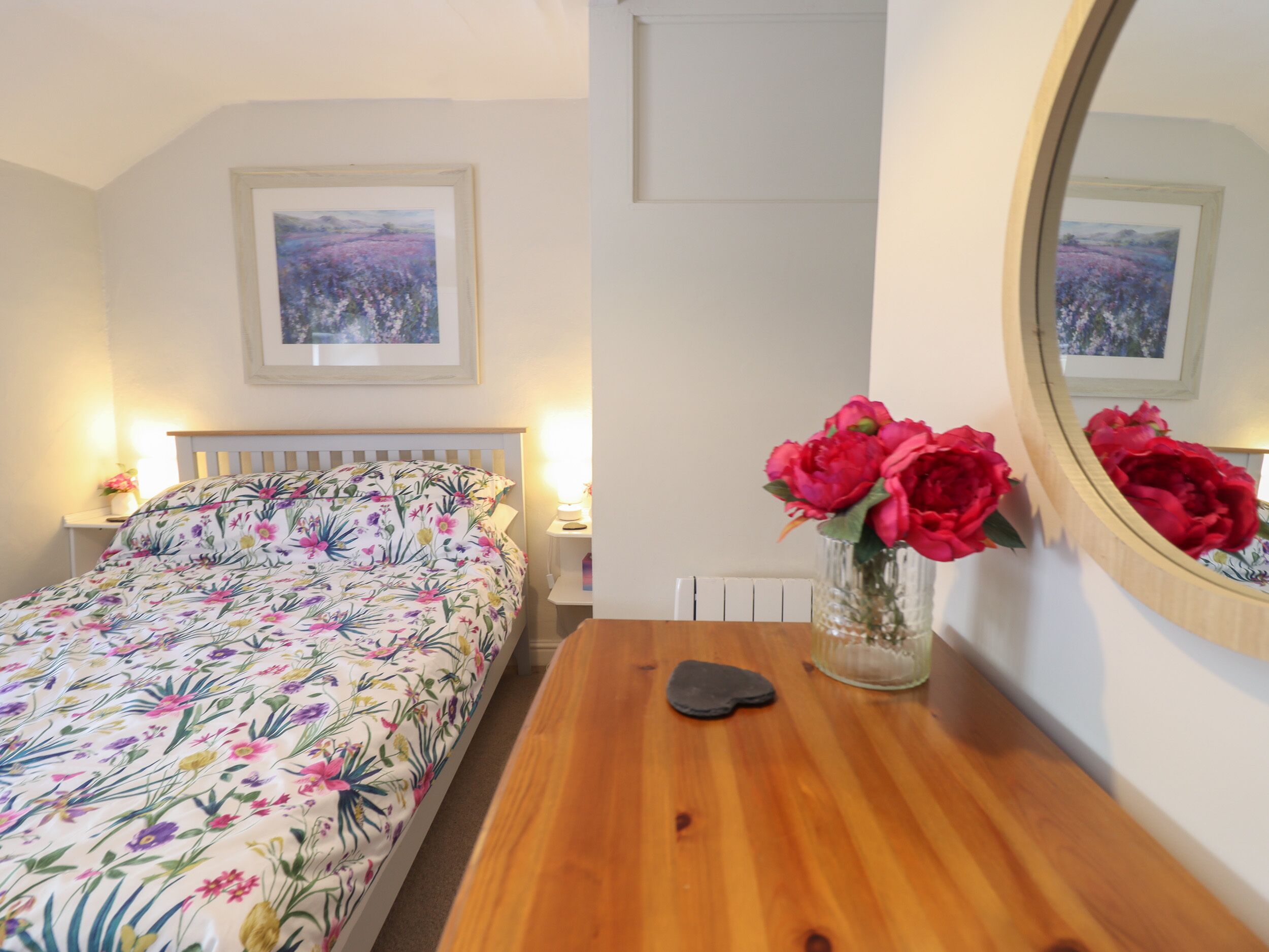 A bedroom with a floral bedspread, a wooden dresser, a vase with red flowers, and a framed picture on the wall.