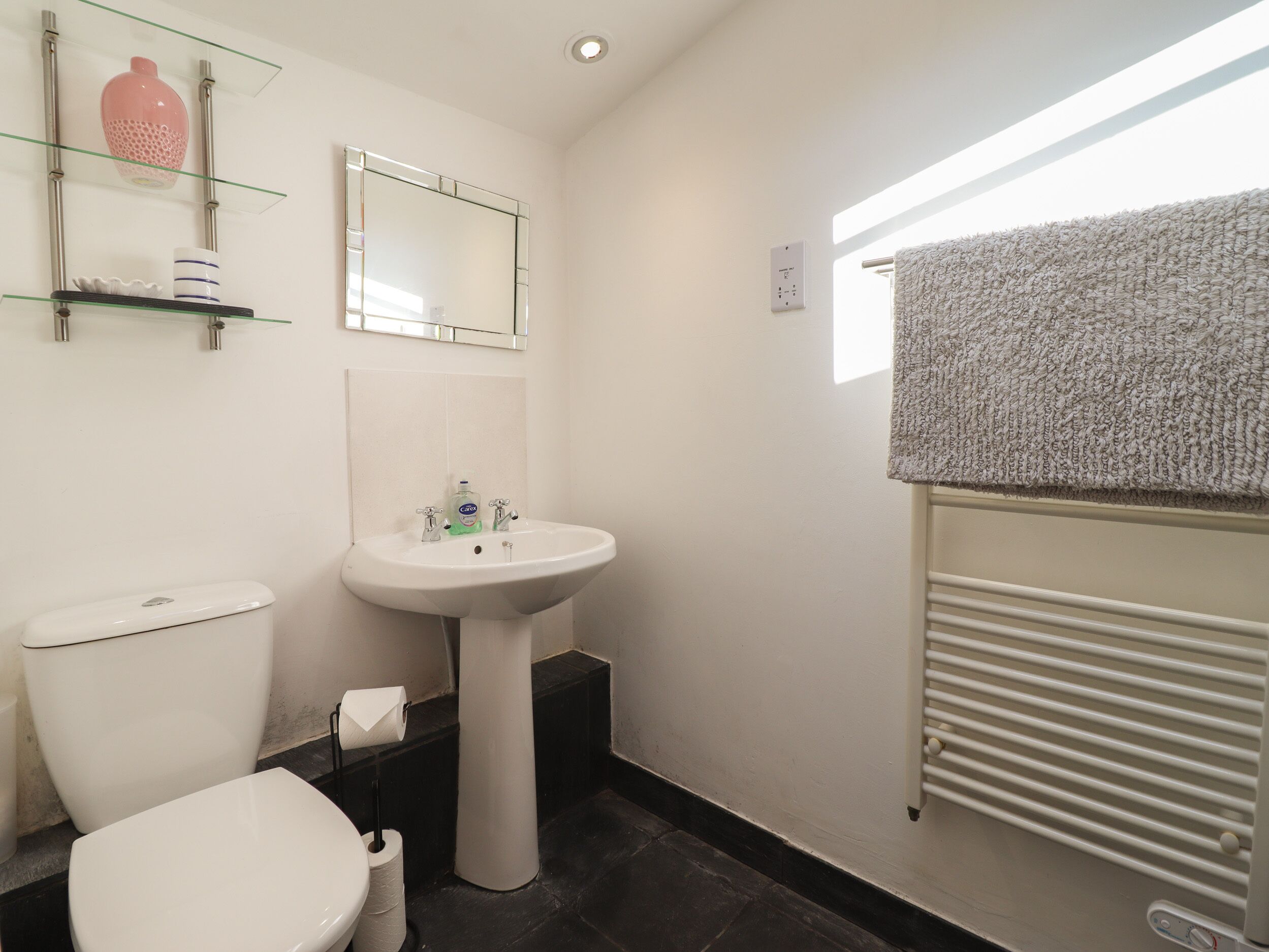 Et badeværelse med toilet, håndvask, et glas hyld med toiletartikler, et håndklædestativ og en radiator.