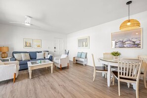 Una sala de estar moderna con un sofá azul, sillones blancos, una mesa de centro de vidrio y un comedor con mesa blanca y sillas de madera.