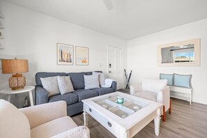 Una sala de estar moderna con un sofá gris, un sillón blanco y una mesa de centro de vidrio. Hay cuadros enmarcados en la pared y una ventana con vistas a un edificio.
