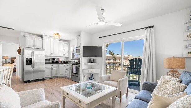 Una cocina moderna con armarios blancos, un refrigerador de acero inoxidable y un televisor de pantalla plana. Hay una zona de comedor con una mesa y sillas blancas, y una sala de estar con un sofá azul y una mesa de centro de vidrio.