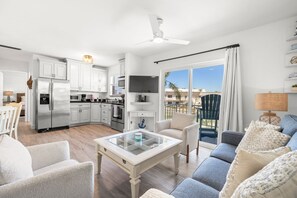 Una cocina moderna con armarios blancos, un refrigerador de acero inoxidable y un televisor de pantalla plana. Hay una zona de comedor con una mesa y sillas blancas, y una sala de estar con un sofá azul y una mesa de centro de vidrio.