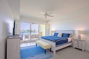 A bedroom with a bed, bedside tables, a bench, a ceiling fan, and a view of the outside.