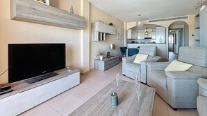 A modern living room with a flat-screen TV, grey sofas, a coffee table, and a kitchen in the background.