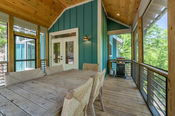 A wooden deck with a dining table and chairs, a grill, and a view of the outdoors.