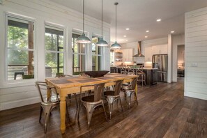 A spacious kitchen with a wooden dining table, metal chairs, pendant lights, and a view of the outdoors through large windows.
