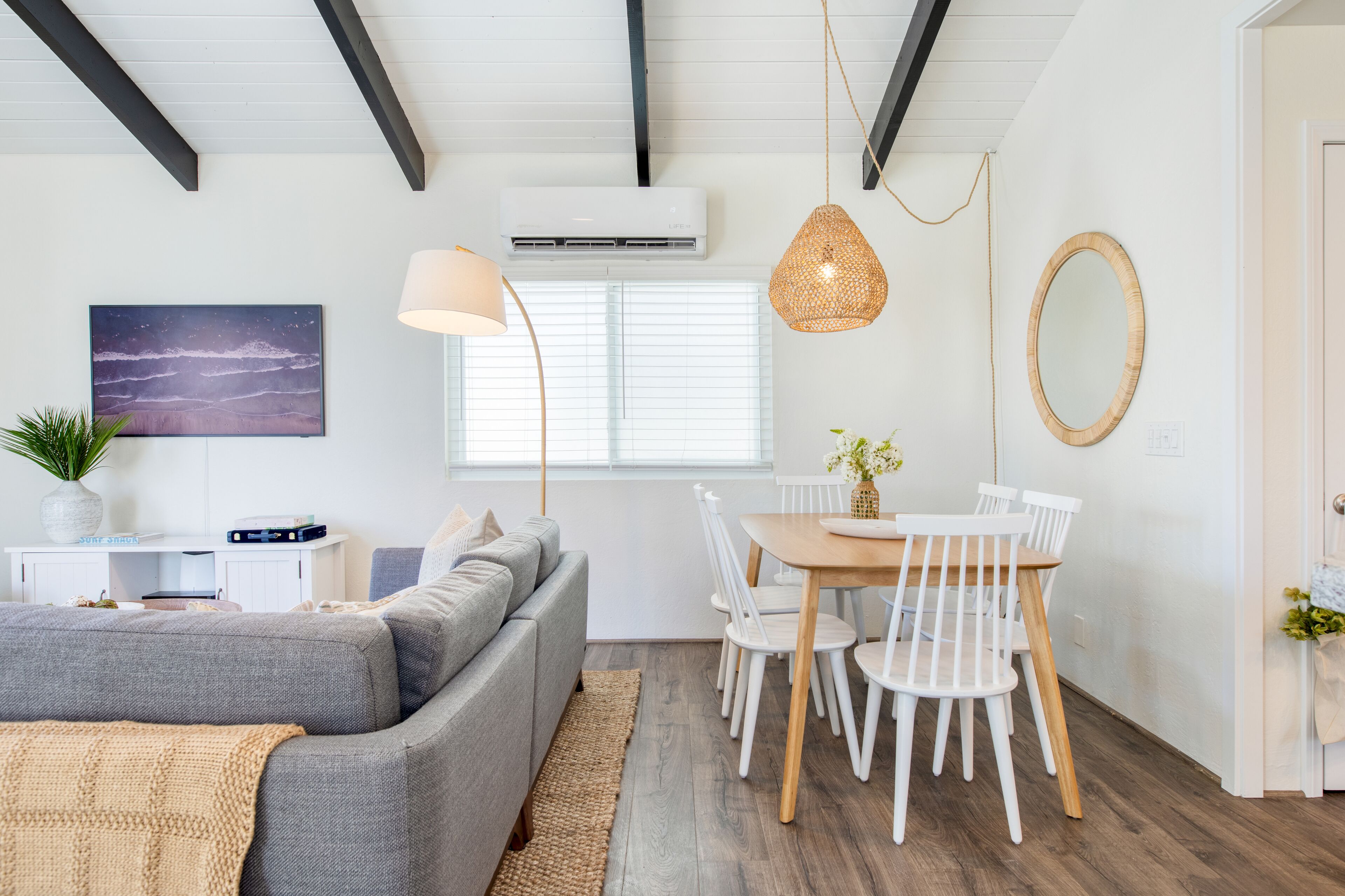 A modern living room with a grey sofa, a dining table with white chairs, a hanging woven light fixture, and a wall-mounted air conditioner.