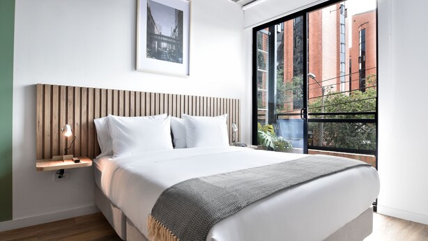 Un dormitorio moderno con una cama grande, un cabecero de madera, una mesita de noche con lámpara, un cuadro enmarcado en la pared y vistas a edificios a través de un ventanal amplio.