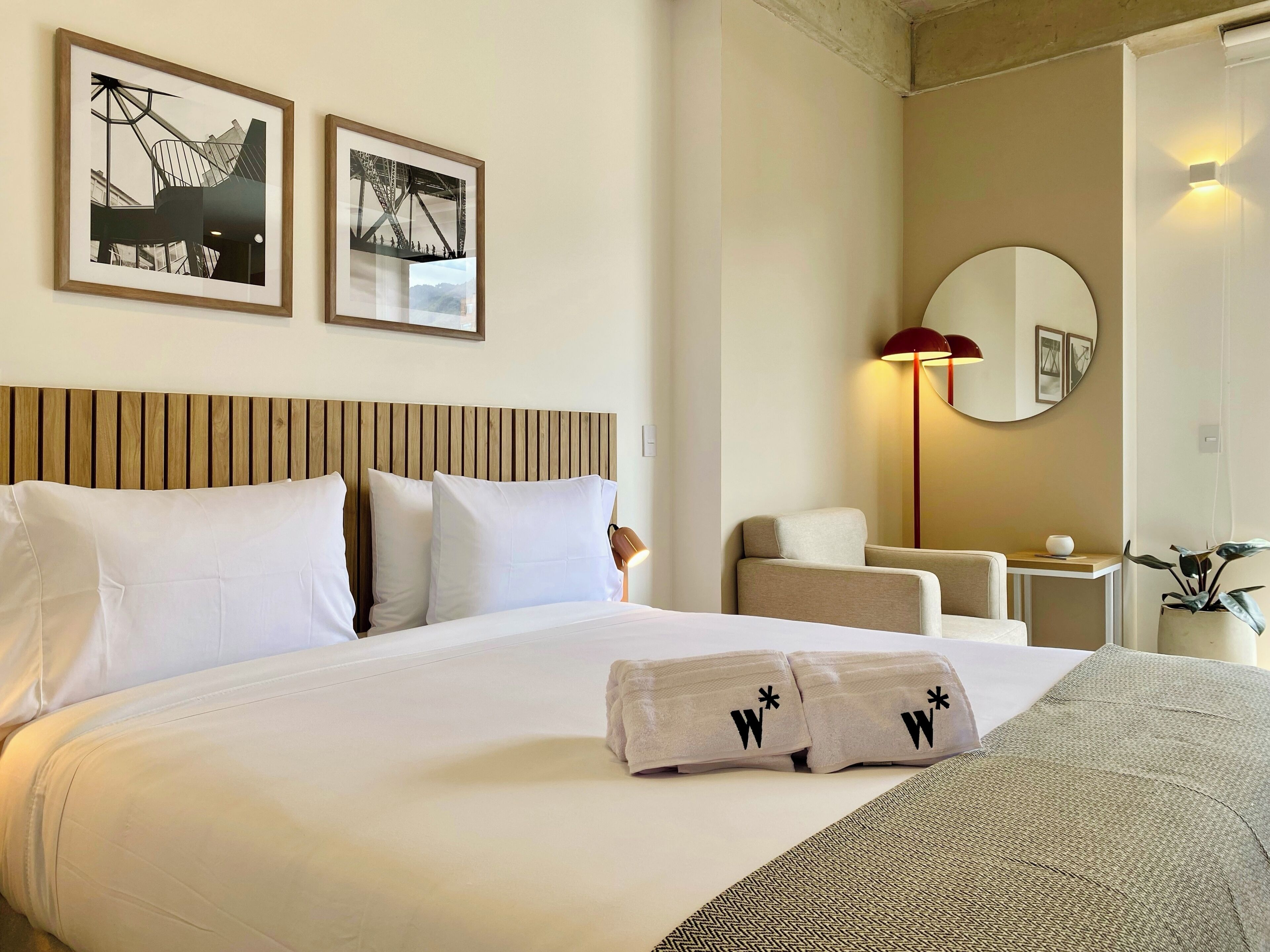 A hotel room with a bed, two framed pictures on the wall, a round mirror, a desk, and a chair.