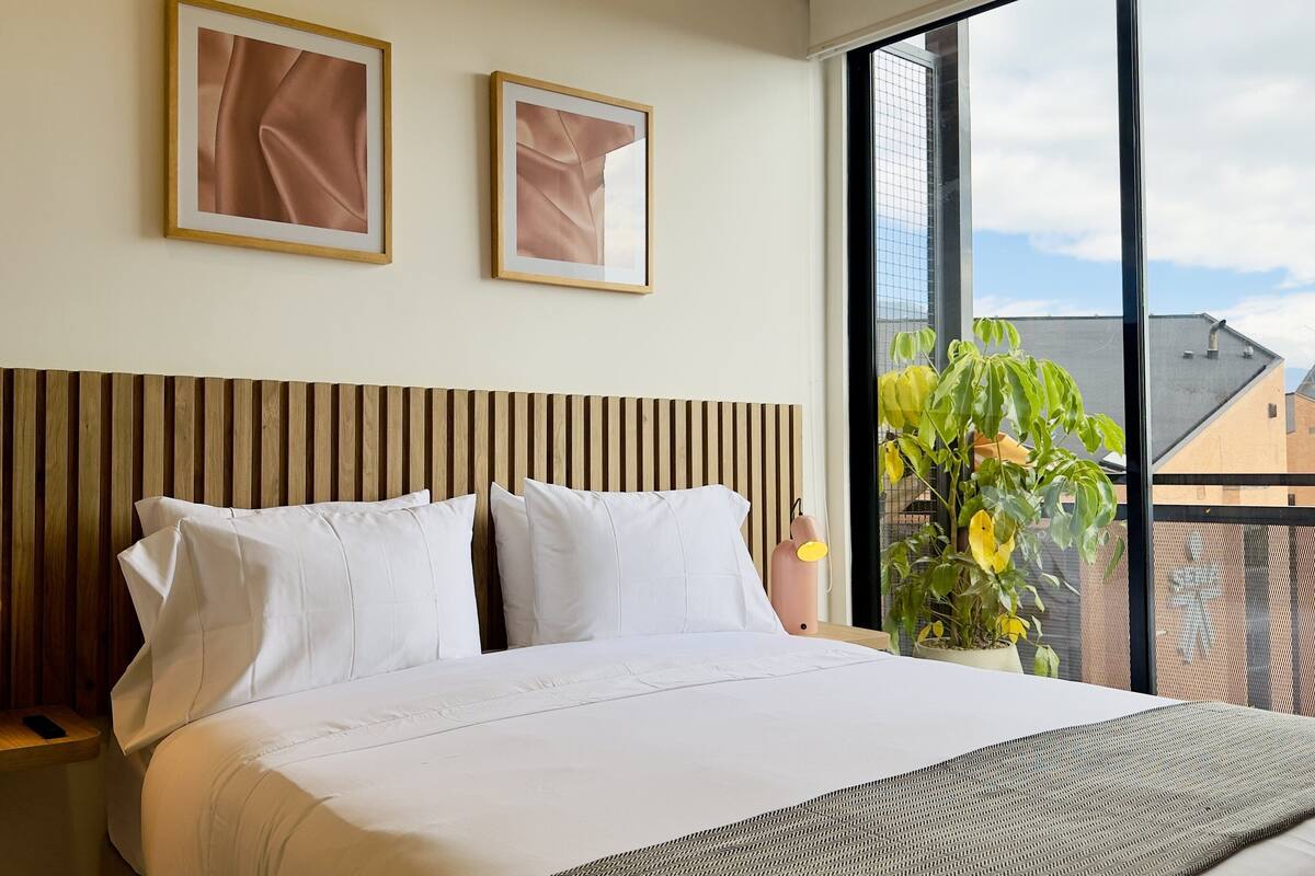 Ein Hotelzimmer mit einem Holzkopfteil, weißer Bettwäsche, zwei gerahmten Bildern an der Wand und Blick auf ein Gebäude durch ein großes Fenster.