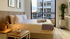 Una habitación de hotel moderna con una cama grande, una mesita de noche con lámpara, una pequeña mesa con una taza, una planta en maceta y vistas a un edificio a través de un ventanal amplio.