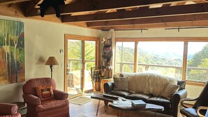 A spacious living room with wooden beams, a leather sofa, a red armchair, and a large window offering a view of the outdoors.