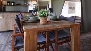 Une table à manger rustique en bois, agrémentée d’un panier tressé en guise de centre de table, est entourée de chaises aux couleurs sombres. On aperçoit en arrière-plan une cuisine avec des meubles en bois et un luminaire suspendu.