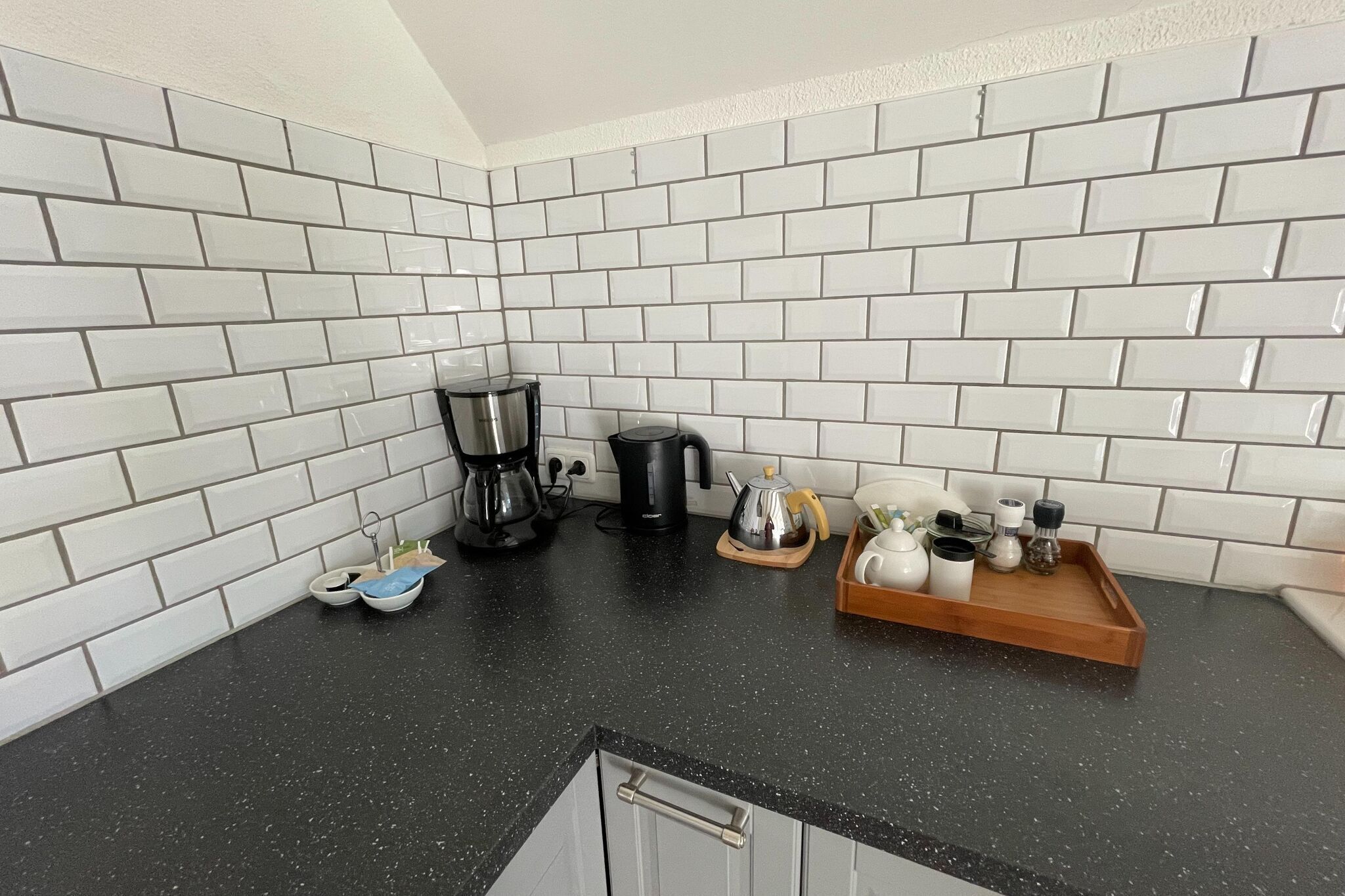 A kitchen with white subway tiles, a black countertop, a coffee maker, a kettle, and a tray with condiments.