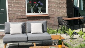 A grey sofa with white cushions, a wooden table with a bottle and glasses, and a window with flowers. - Vacation home in Schoonloo with terrace (Schoonloo)