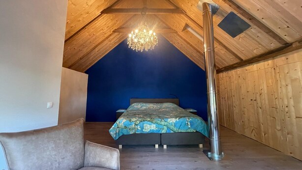 A bedroom with a sloped ceiling, a chandelier, a bed with a floral bedspread, and a tall, cylindrical object on the floor. - Vacation home in Schoonloo with terrace (Schoonloo)