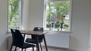 A dining area with a table and chairs, a window with a view of trees and flowers, and a radiator below the window. - Vacation home in Schoonloo with terrace (Schoonloo)