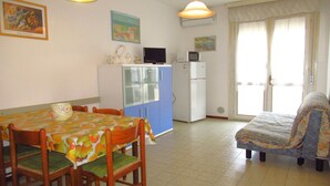 A dining area with a table and chairs, a kitchenette with a refrigerator and microwave, and a sofa in a living area with a view through a window. - Apartment in Caorle (VE) (Caorle (VE))