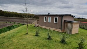 A small, single-story wooden cabin with a porch, surrounded by a well-maintained lawn and a field in the background.