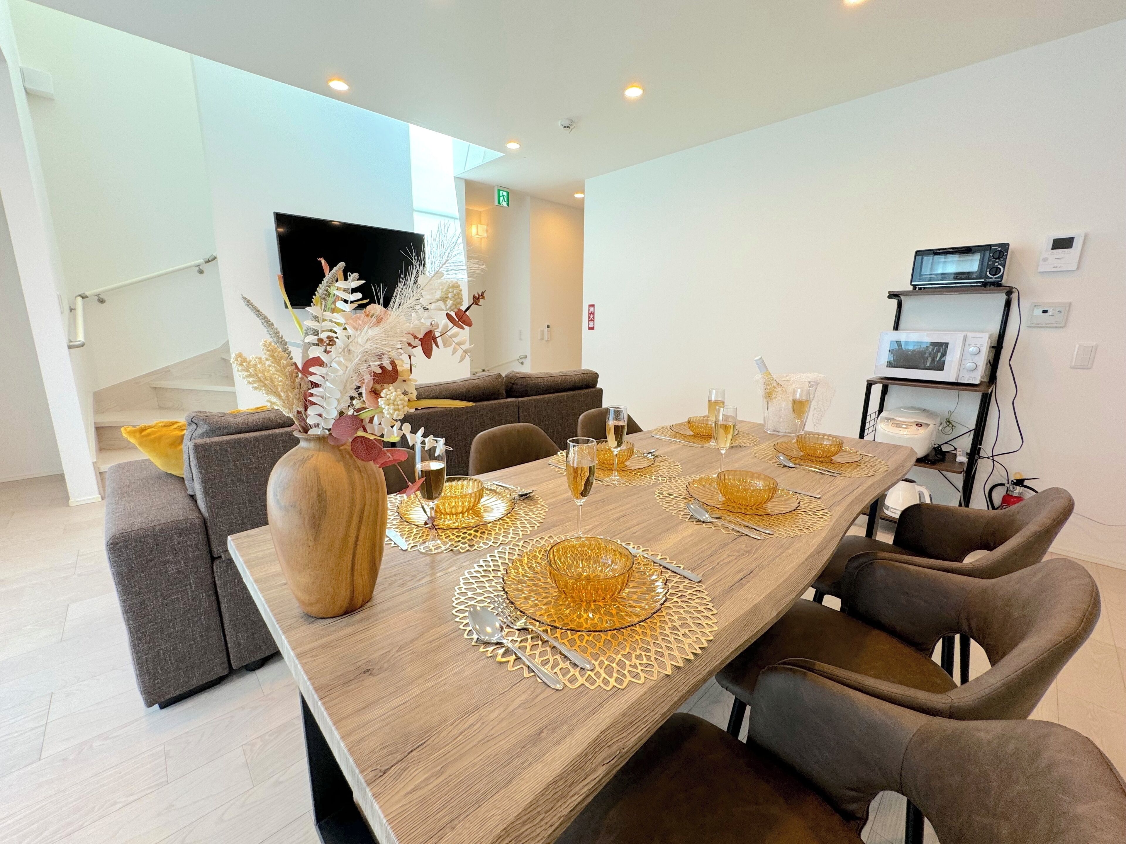 Un espace repas moderne avec une longue table en bois, dressée pour six personnes, entourée de chaises à revêtement gris. Un vase contenant des fleurs séchées est posé sur la table, et un téléviseur à écran plat est fixé au mur.