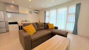 A modern living room with a brown sectional sofa, a wooden coffee table, and a kitchen area in the background. - Grandioso Okinawa Pool Villa Kin 2 (Kin)