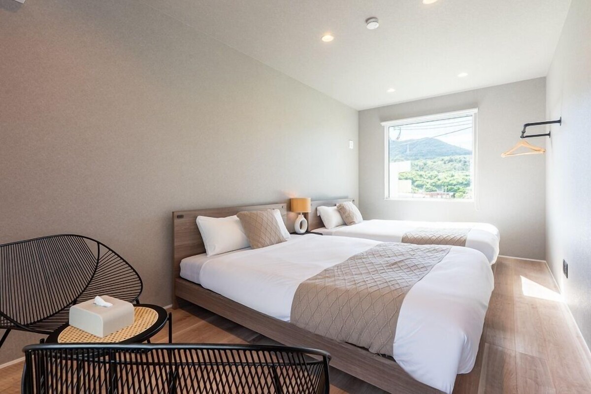 A modern hotel room with two beds, a sitting area with a chair, a small table, and a window with a view of greenery.